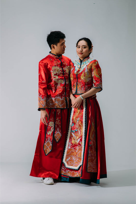 Bride and groom in matching traditional red and gold Chinese wedding outfits, standing together with intricate embroidery details, set against a neutral background.