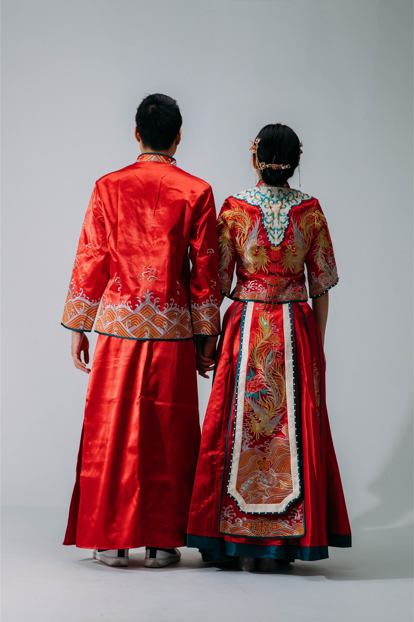 Rear view of bride and groom in matching traditional red and gold Chinese wedding outfits, showcasing intricate embroidery details, set against a neutral background.