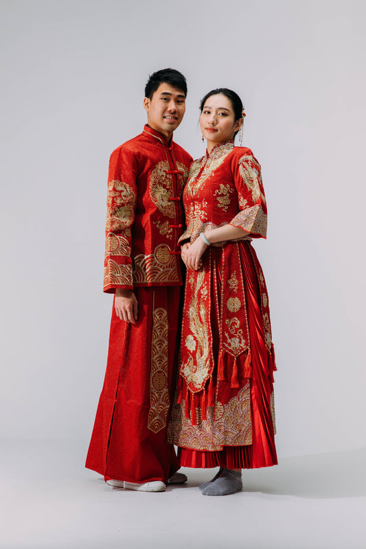 Bride and groom wearing matching traditional red Chinese wedding outfits with intricate gold embroidery, standing together against a neutral background.