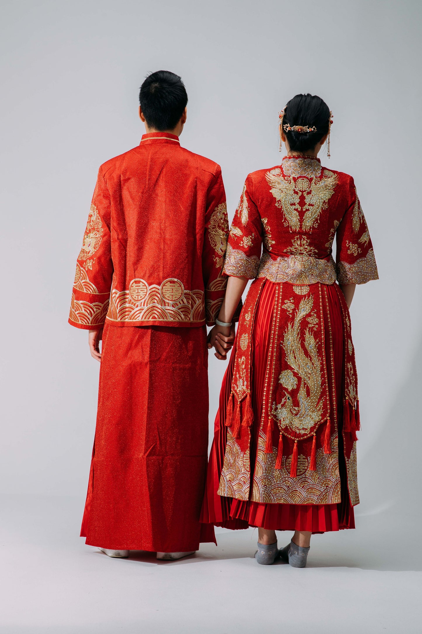 Rear view of bride and groom wearing matching traditional red Chinese wedding outfits with intricate gold embroidery, standing together against a neutral background.