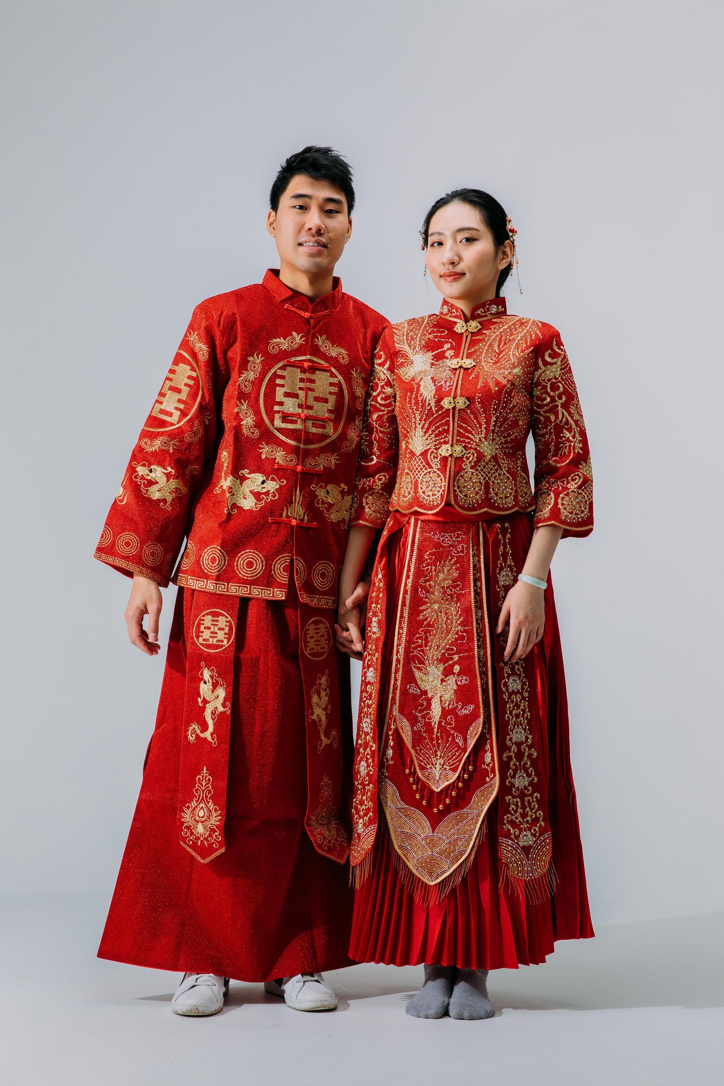 Bride and groom dressed in matching traditional red and gold Chinese wedding outfits with intricate dragon and phoenix embroidery, standing against a neutral background.