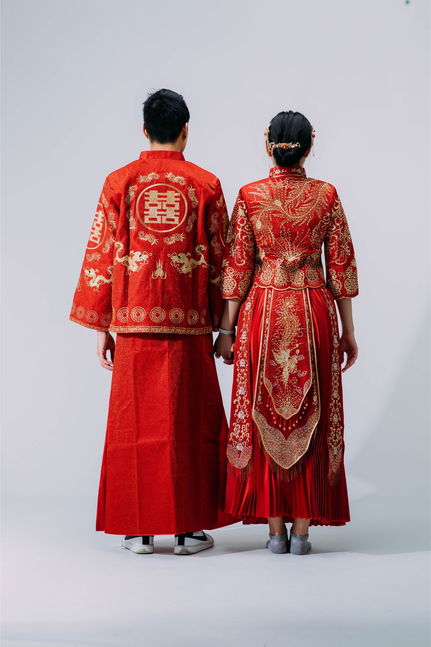 Rear view of bride and groom wearing traditional red and gold Chinese wedding outfits with detailed embroidery of dragon, phoenix, and Double Happiness motifs, standing against a neutral background.