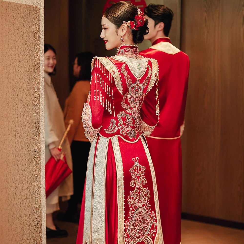 Bride in Imperial Phoenix traditional Chinese wedding dress, back view showing gold embroidery and tassel detail, groom and guests in background.