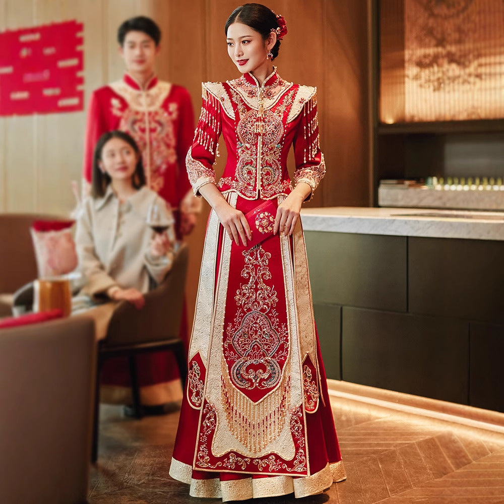 Bride in Imperial Phoenix traditional Chinese wedding dress featuring ornate gold embroidery and shoulder tassels, standing in a modern venue with groom and guests in background.